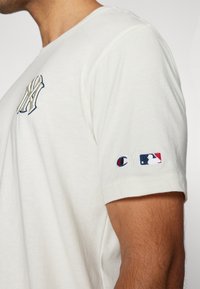 Kremkrāsas kokvilnas t-krekls ar izšūtu New York Yankees logo uz krūtīm un mazu Champion/MLB logo uz piedurknes. Īsas piedurknes.