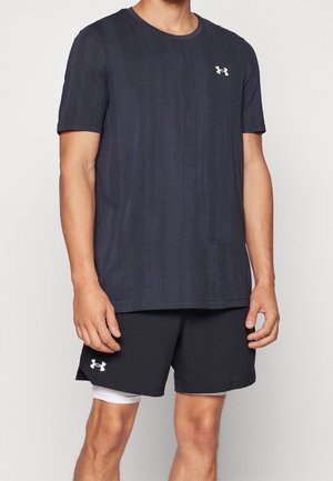 Mężczyzna ubrany w ciemną koszulkę Under Armour z krótkim rękawem i pasujące krótkie spodenki z białymi spodenek kompresyjnych pod spodem, stojący na białym tle.