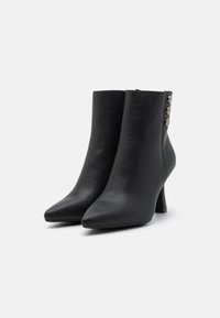 Botas de tobillo de cuero negro con puntas afiladas, tacones stiletto medianos y tres botones decorativos dorados en el costado. Textura suave.