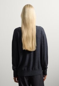 Persona con lunghi capelli biondi lisci, che indossa un maglione grigio scuro e pantaloni neri, in piedi di fronte a un muro bianco semplice.