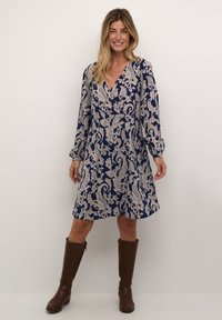 Robe marine avec un motif paisley crème, en v, à manches longues et taille froncée. Des bottes brunes jusqu'aux genoux complètent la tenue.