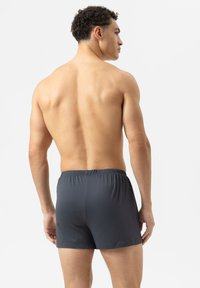 Un homme torse nu vu de dos, portant un short texturé gris foncé, se tenant devant un fond blanc uni.