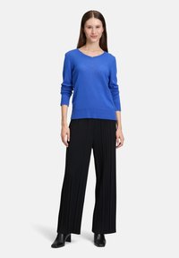 Femme debout portant un pull bleu à manches longues, un pantalon large plissé noir et des bottines noires sur un fond blanc uni.