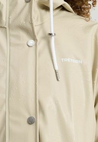 Beige vedenpitävä takki, jossa on hopeiset nepparit, valkoinen kiristysnarun ja brändilogo "Tretorn" näkyvissä. Pinnassa näkyy vesipisaroita.