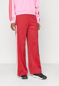 Piros, bő szabású nadrág puha anyagból, fehér oldalsávokkal. Az egyén fekete sportcipőt és rózsaszín pulóvert visel.