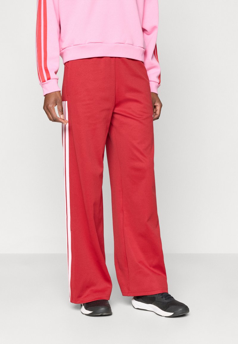 Piros, bő szabású nadrág puha anyagból, fehér oldalsávokkal. Az egyén fekete sportcipőt és rózsaszín pulóvert visel.