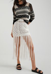 Černobílý pletený svetr s vzorem a průhledná bílá háčkovaná sukně s dlouhými třásněmi, doplněné černými nazouváky.