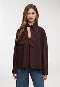 Blusa marrone con collo alto arricchito da grandi decorazioni a forma di foglia e apertura a goccia, realizzata in tessuto liscio con maniche lunghe.