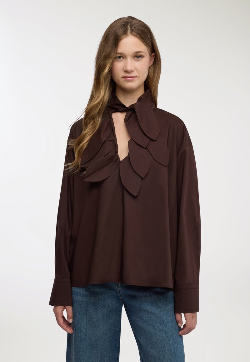 Blusa marrone con collo alto arricchito da grandi decorazioni a forma di foglia e apertura a goccia, realizzata in tessuto liscio con maniche lunghe.