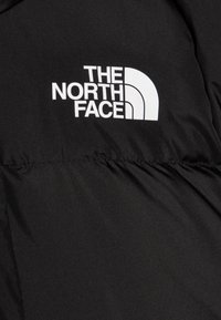 Juodas pūkinis švarkas su ryškiu baltu "The North Face" logotipu ant krūtinės, su lygaus paviršiaus tekstūra ir siuvinėtais akcentais.
