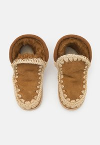 Chaussons en daim marron avec doublure en fourrure, présentant des accents beiges tressés et un bout arrondi. La texture douce et les détails cousus améliorent le design.