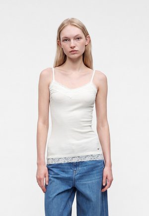 Jeune femme aux longs cheveux blonds, portant un caraco blanc bordé de dentelle et un jean bleu ample, debout devant un fond uni.