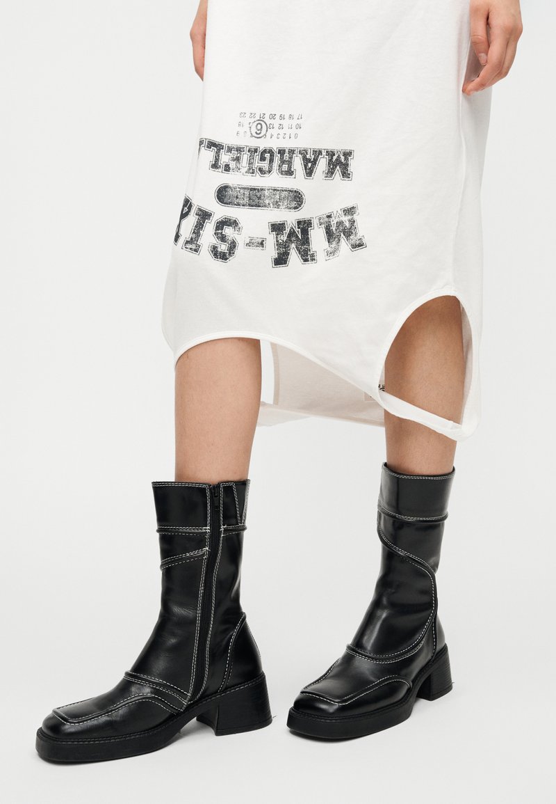 Bottes en cuir noir jusqu'à la cheville avec un design semi-structuré, surpiqûres blanches en contraste, bout carré et talon bloc. Fermeture éclair latérale pour un enfilage facile.