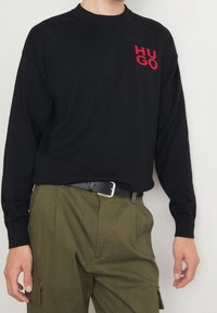 Pull en maille noire avec un col rond et un logo rouge "HUGO". Associé à un pantalon vert et une ceinture noire avec une boucle argentée.