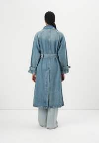 Lyseblå denimjakke med belte i midjen, lange ermer og oppbrettede mansjetter. Bakfra vises en rett snitt og midtlange design.