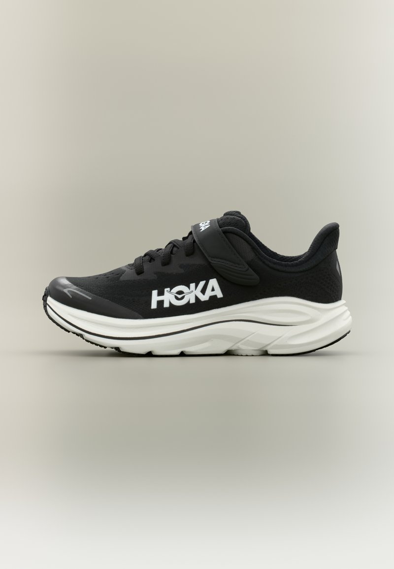Μαύρο αθλητικό παπούτσι με λευκή σόλα, κορδόνια και λουράκι, με το λογότυπο "HOKA" στο πλάι.