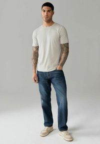 T-shirt grigio chiaro a maniche corte con scollo rotondo, abbinato a jeans in denim blu e sneakers beige. Tatuaggi visibili sulle braccia.