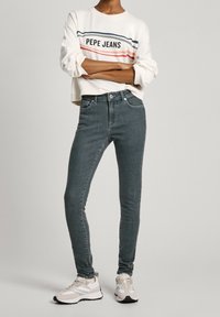 Světle šedé skinny džíny s hladkou texturou, kombinované s bílou mikinou s barevným pruhovaným designem. Doplňují je tenisky.