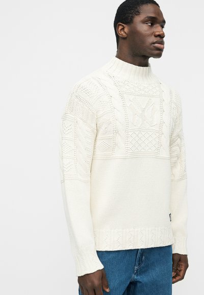 Άνδρας που φοράει λευκό πλεκτό πουλόβερ με πλεξούδες και ψηλό γιακά, συνδυασμένο με μπλε τζιν, στραμμένος προς τα δεξιά σε απλό φόντο.