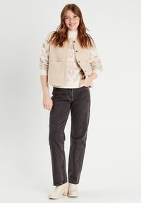 Gilet en polaire beige avec une surface texturée sur un pull blanc présentant un motif rose doux, associé à un jean noir et des bottes couleur crème.