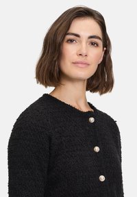 Schwarze, strukturierte Strickjacke mit rundem Halsausschnitt, ausgestattet mit drei Perlenknöpfen. Der Stoff hat ein weiches, flauschiges Aussehen.