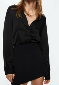 Blusa di seta nera con bottoni e scollatura a V, abbinata a una gonna nera. Tessuto liscio, maniche lunghe e vestibilità sartoriale.