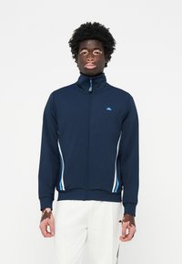 Mornarsko modra zip-up jakna z visokim ovratnikom, ki ima bel in svetlo moder stranski trak. Izdelana iz gladkega, raztegljivega materiala. Modra logotip na prsih.