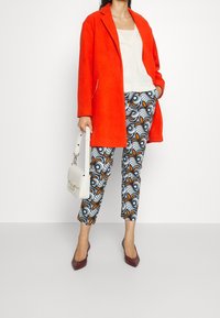 Donna che indossa un cappotto arancione brillante, top bianco, pantaloni corti a fantasia, tacchi bordeaux, che tiene una borsa bianca, in piedi davanti a uno sfondo semplice.