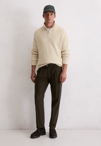 Beige geribbelde trui met een ritskraag, gecombineerd met donkergroene broek en donkere schoenen. Het model draagt een bijpassende pet.