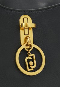 Primo piano di una borsa a tracolla in pelle nera con un fermo in metallo dorato e un ciondolo circolare con dettagli del logo. Texture liscia e design minimalista.