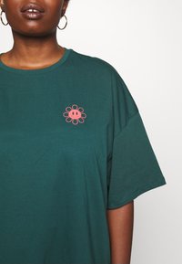 Groene katoenen t-shirt met een ontspannen pasvorm, voorzien van een roze bloemenprint op de linkerborst en korte mouwen.