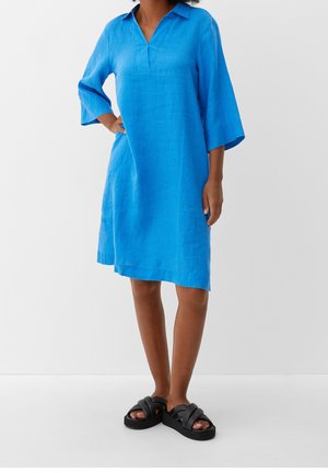 Woman wearing a knee-length, bright blue linen dress with 3/4 sleeves and black platform sandals, standing with one hand on hip.