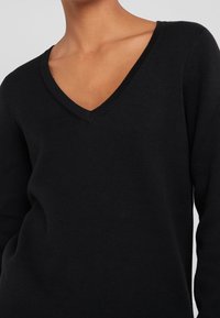 Pull en tricot noir avec un décolleté en V, doté de manches longues et d'une texture douce. Le tissu semble légèrement côtelé et léger.