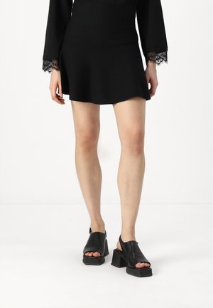 Robe noire courte avec des accents en dentelle sur les manches, assortie à des sandales noires à talons bloc ouvertes, mettant en valeur une texture en cuir lisse.