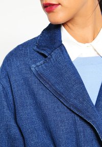 Niebieska dżinsowa marynarka z wyciętym kołnierzem, z teksturowanym materiałem i widocznymi detalami ściegów. Nałożona na jasnoniebieski sweter.