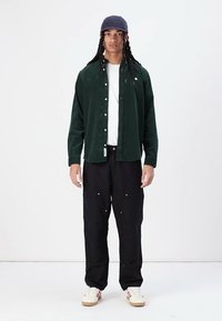 Cămașă verde din fustă cu butoni peste un tricou alb, asortată cu pantaloni cargo negri largi cu buzunare și adidași albi cu dungi roșii.
