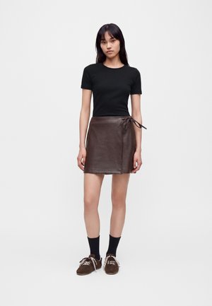 Femme debout portant un haut noir à manches courtes, une jupe portefeuille en cuir marron avec un lien sur le côté, des chaussettes noires et des chaussures marron à lacets sur fond blanc.