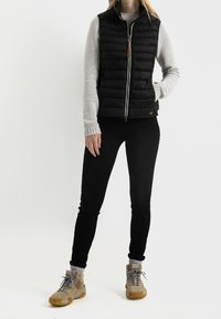 Svart quiltad väst med hög krage och dragkedja, kombinerad med en ljusgrå tröja, svarta jeans och beige vandringskängor.