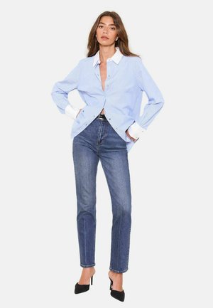 Woman wearing a loose light blue striped shirt with white cuffs and collar, high-waisted blue jeans, and black pointed heels, standing with hands in pockets.