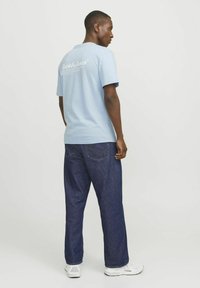 Camiseta de manga corta azul claro con cuello redondo y logo en la parte posterior. Combinada con unos vaqueros anchos azul oscuro y zapatillas blancas.