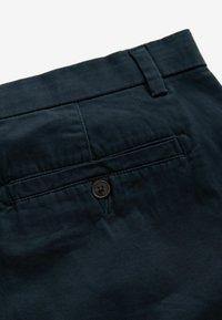 Dunkelblaue Baumwollhosen mit einer Gesäßtasche, die einen braunen Knopf und gestickte Details aufweist. Strukturierter Stoff mit glatter Oberfläche.