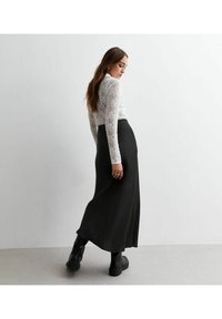 Zwarte maxi rok met een gladde, vloeiende textuur en hoge taille, gecombineerd met een strakke witte kanten top met lange mouwen. Zwarte laarzen maken de look compleet.