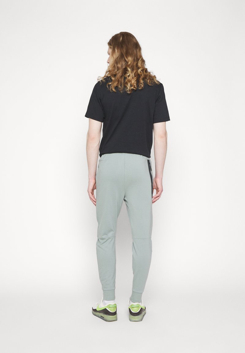 Joggings vert clair avec des jambes effilées, t-shirt noir, et sneakers blanches avec des accents verts, vus de derrière sur un fond uni.