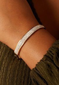 Goudkleurige armband met een rij heldere kristallen in het midden, afgezet tegen een getextureerde, warme huidtint.