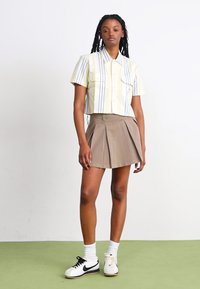 Camicetta corta a maniche corte a righe giallo chiaro con due tasche frontali, abbinata a una mini gonna plissettata color cammello e scarpe da ginnastica nere e bianche.