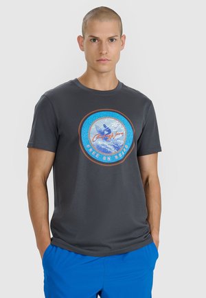 Mężczyzna z krótkimi włosami, ubrany w ciemnoszary t-shirt z grafiką przedstawiającą niebieski okrągły motyw surfera oraz jasnoniebieskie krótkie spodenki, stoi z rękami w kieszeniach.