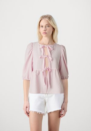 Helles rosa Kurzarm-Blusenshirt mit Schlüsselspaltöffnung, verziert mit zwei Schleifen. Strukturierter Stoff und lockere Passform, kombiniert mit weißen, ausgefransten Shorts.