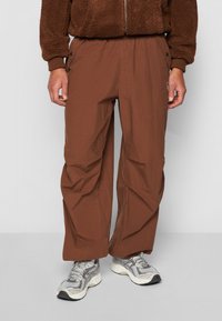 Brune, lette, tætsiddende bukser med sidelommer og en afslappet pasform. Stoffet har en glat tekstur med subtile folder.