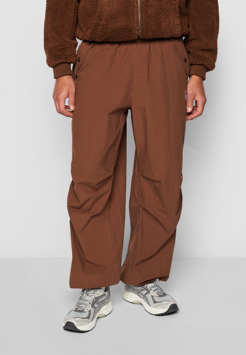 Brune, lette, tætsiddende bukser med sidelommer og en afslappet pasform. Stoffet har en glat tekstur med subtile folder.