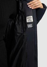 Roka atver melnu jaku, atklājot divas auduma birkas iekšpusē, viena ar tekstu “Siltas apskāviens no auduma,” otra ar zīmola tekstu.